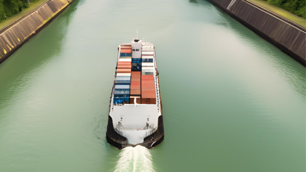 A cargo ship with multiple shipping containers navigates through a canal with green water, flanked by steep embankments on both sides.